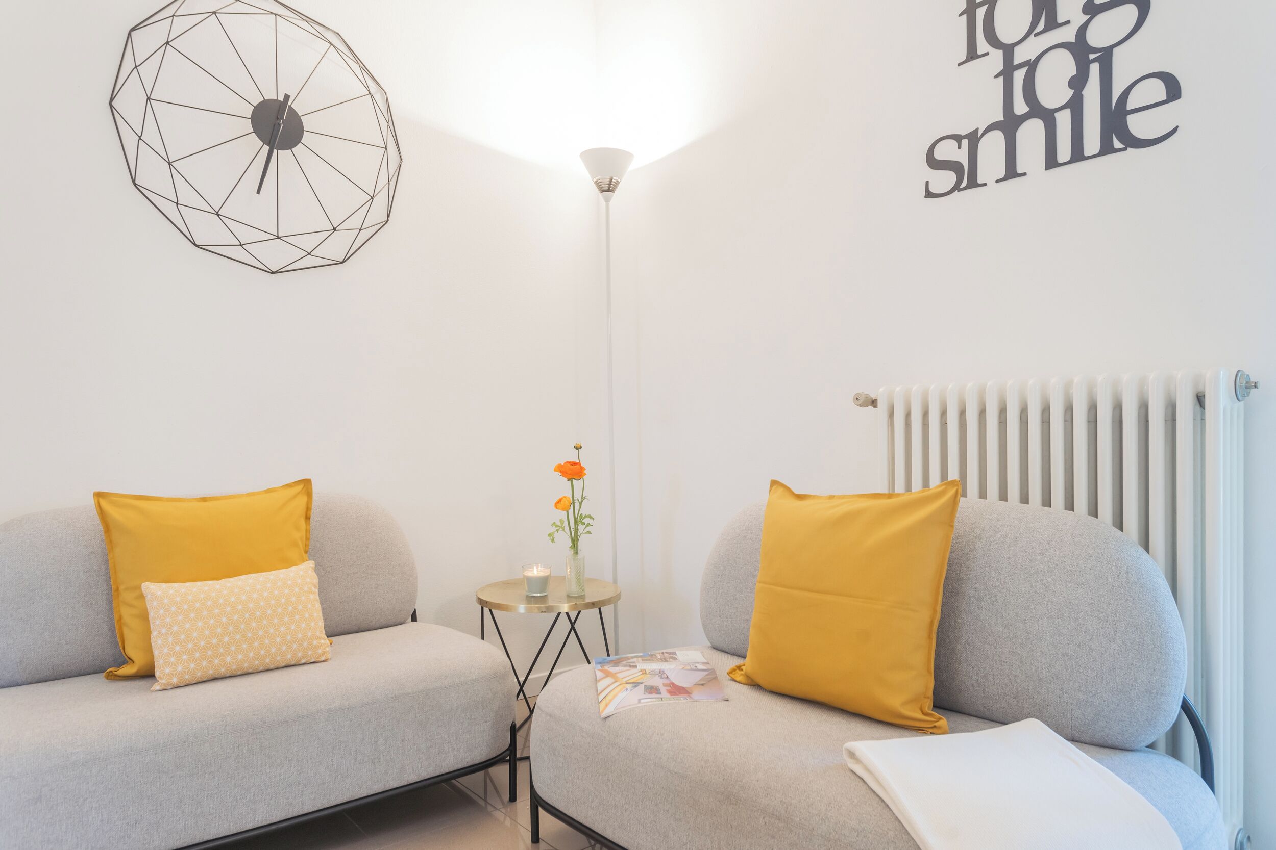 A modern living room with a grey sofa, yellow cushions, a small table with a candle and flowers, and a wall clock.