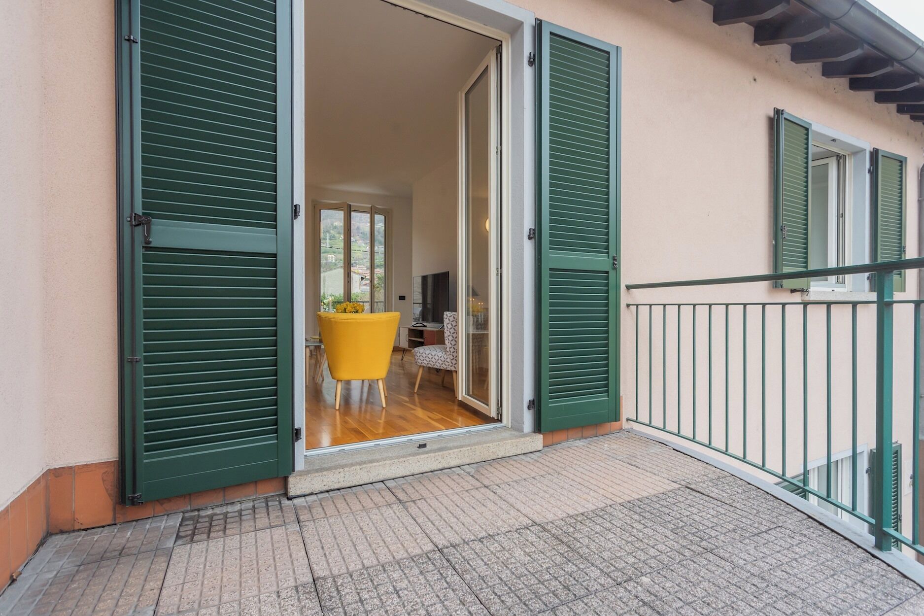 Ein Balkon mit grünen Fensterläden, einem gelben Stuhl und einem Tisch mit einem Blumenstrauß.