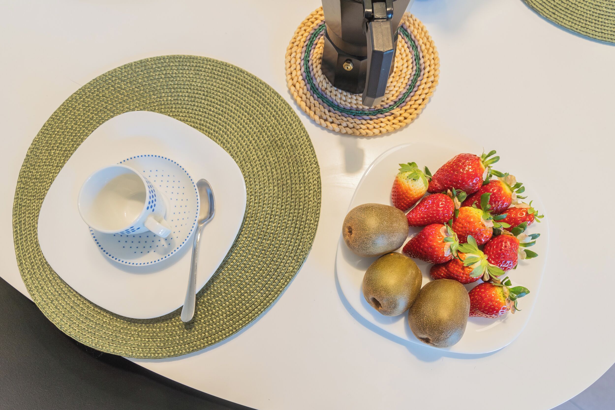A table with a white plate of strawberries and kiwis, a blue cup, and a metal spoon.