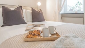 Una cama bien hecha con almohadas, una mesita de noche con una lámpara y una bandeja con pasteles y tazas.