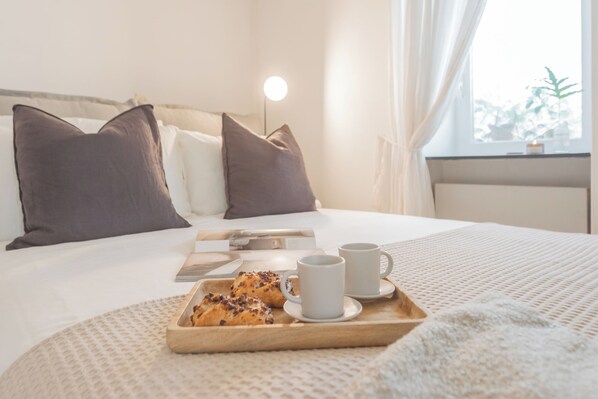 A neatly made bed with pillows, a bedside table with a lamp, and a tray with pastries and cups.
