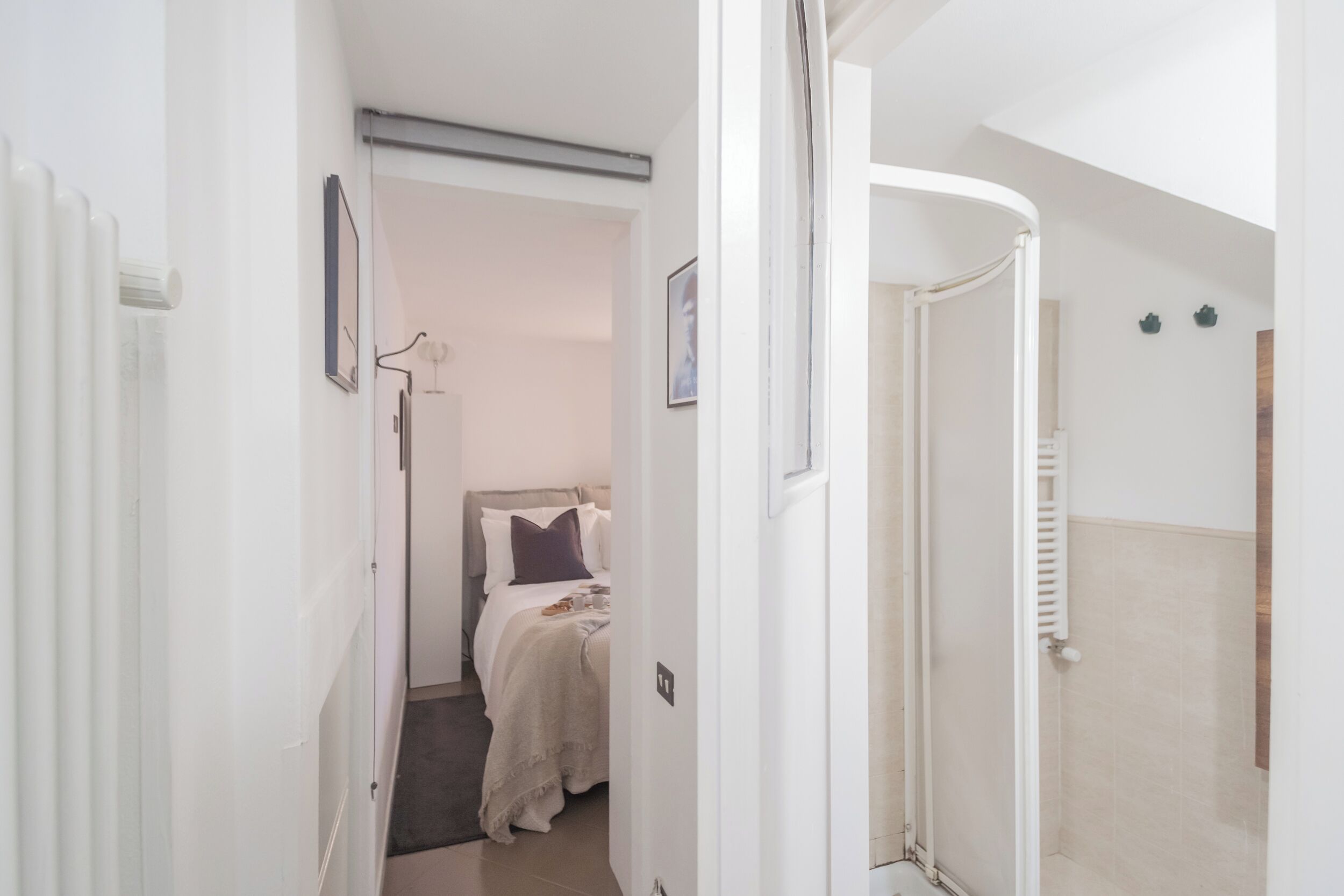 Ein schmaler Flur führt zu einem Schlafzimmer mit Bett, Heizkörper und einer Badezimmertür.