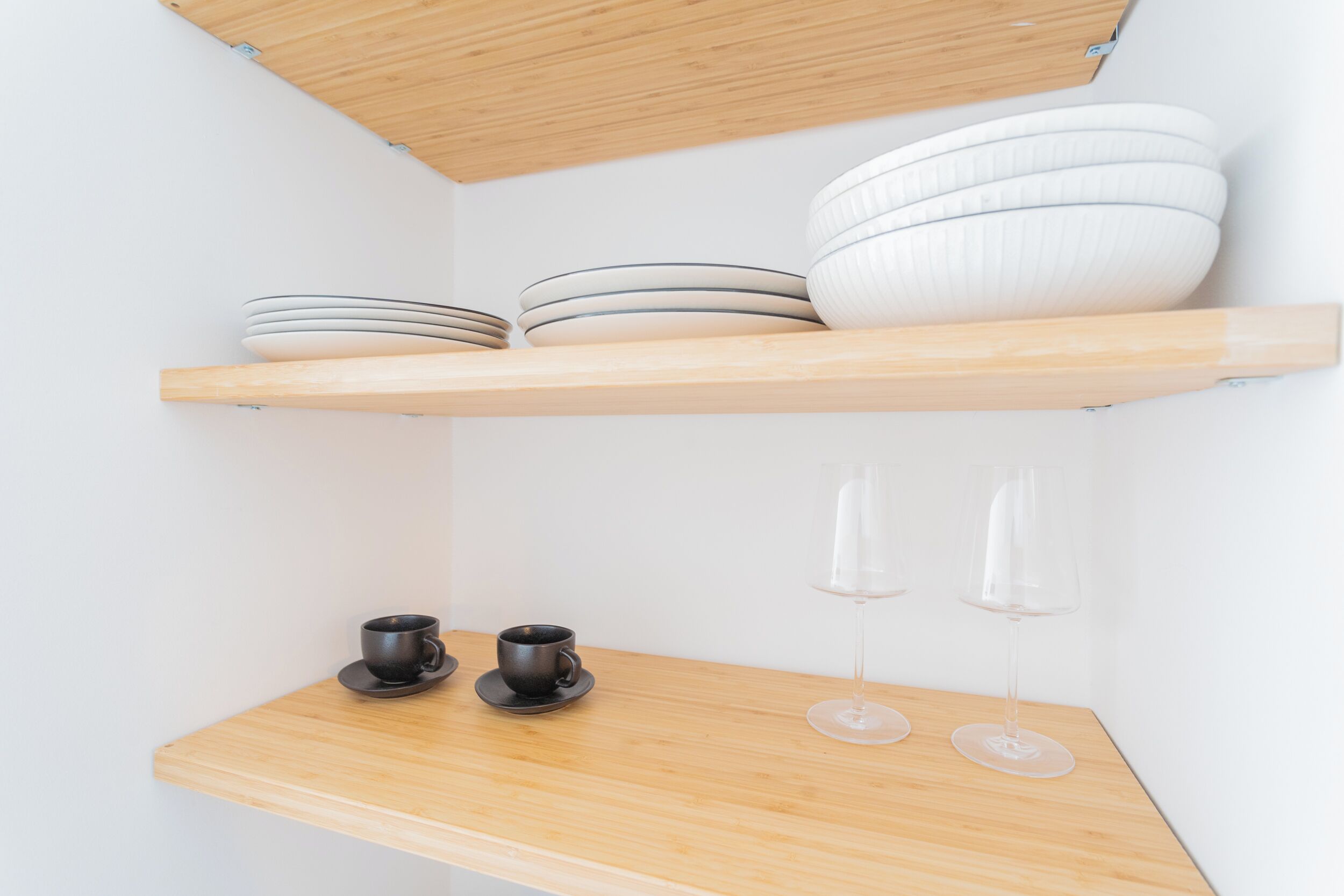 Een minimalistische keuken met houten planken met borden en kommen, en een onderste plank met twee bekers en twee wijnglazen.