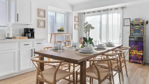 A dining area with a wooden table set for four, surrounded by wicker chairs. A coffee machine and a small plant are on the counter.