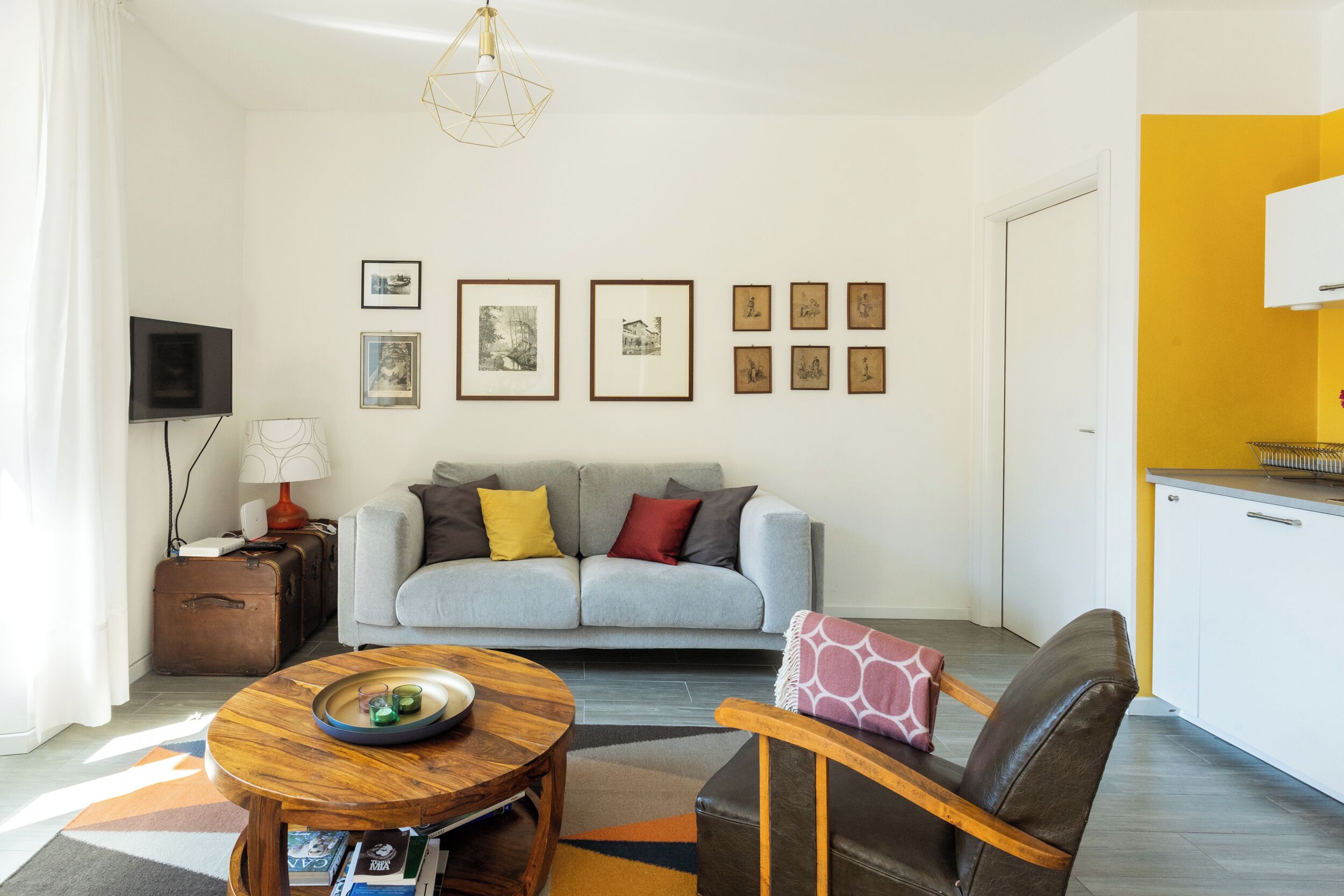 A modern living room with a grey sofa, a wooden coffee table, and a leather armchair. There are framed pictures on the wall and a flat-screen TV on the left.