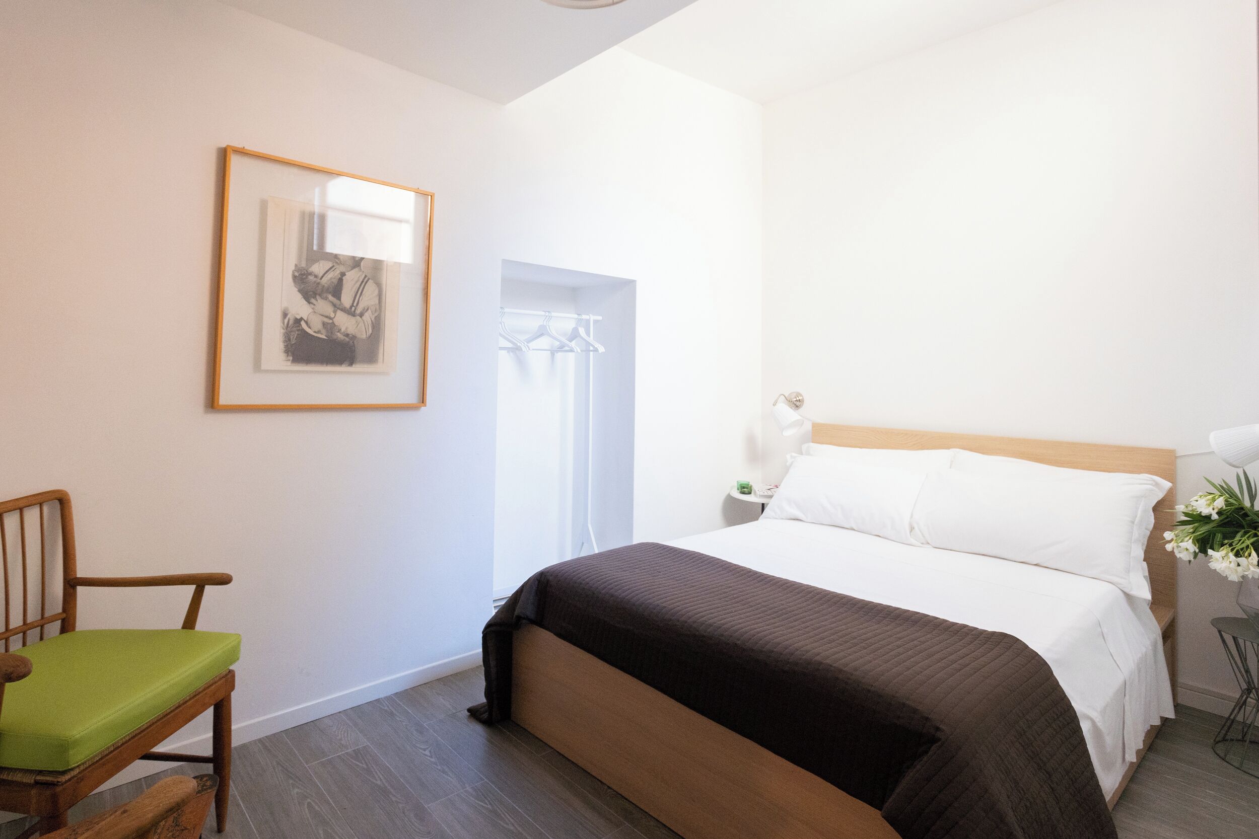 A bedroom with a bed, a chair, a framed picture on the wall, and a nightstand with a lamp.