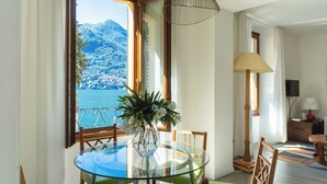 Un comedor con mesa de vidrio, sillas, un jarrón con flores, vista a la montaña y al mar, y una lámpara.