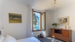 Un dormitorio con cama, cómoda de madera, un cuadro y ventana con vista a una montaña y una masa de agua.