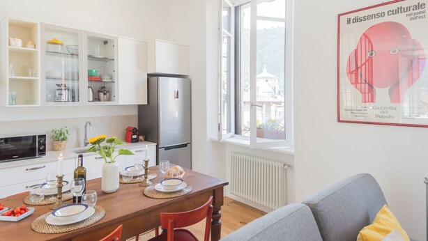 Ett modernt kök med vita skåp, en inbyggd ugn och ett kylskåp. Det finns ett matbord dukat med tallrikar, glas och en vas med blommor. Ett fönster ger utsikt över en byggnad utanför.
