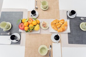 Une table dressée pour le petit-déjeuner, avec deux assiettes de fruits et de viennoiseries, le tout accompagné de tasses et de cuillères assorties.