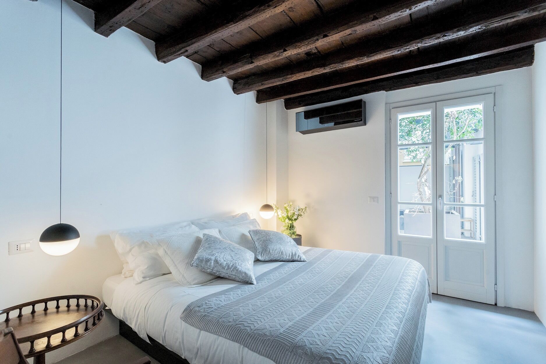 A bedroom with a bed, a bedside table, a hanging light, a window with a view, and a wooden ceiling beam.