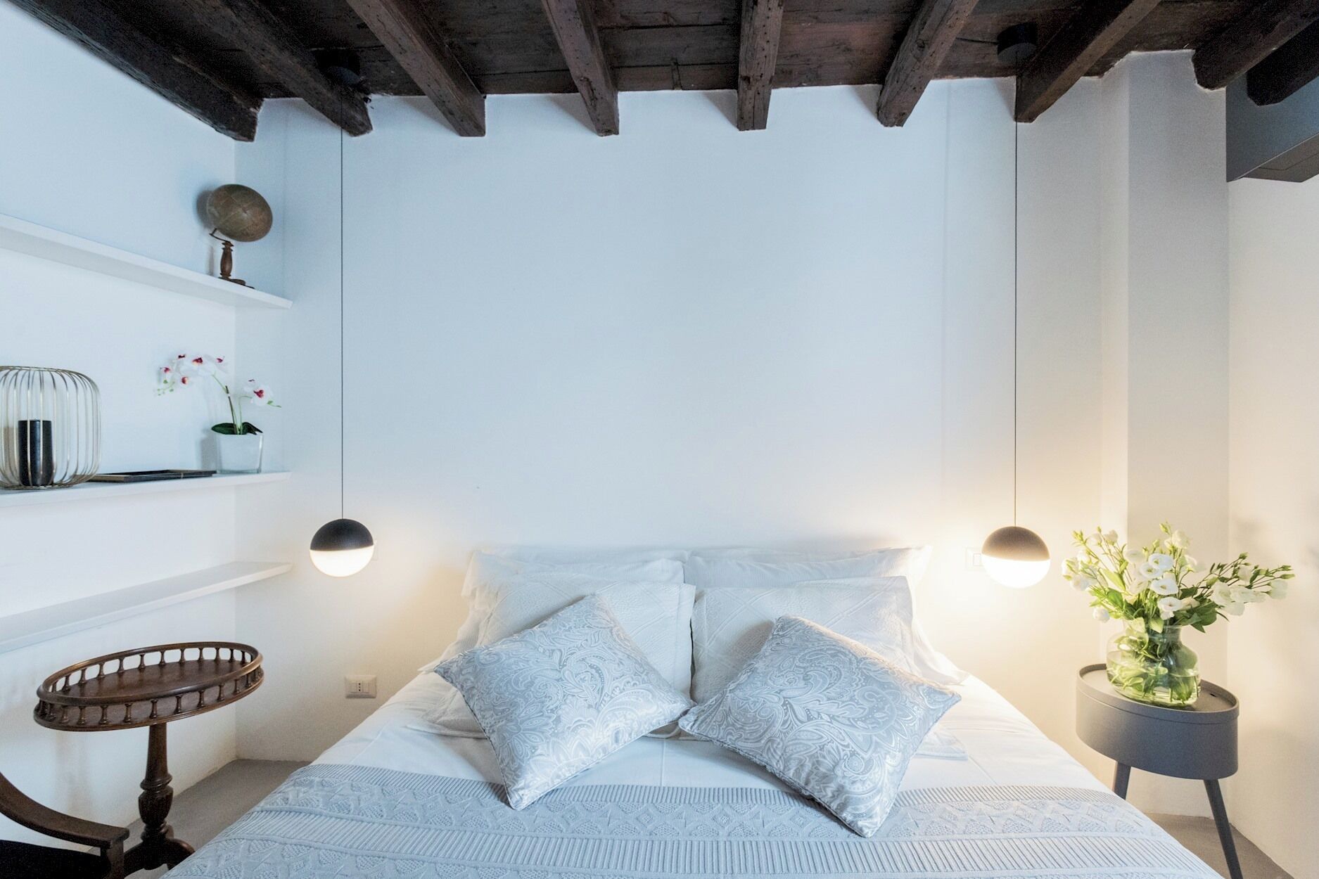 A bedroom with a bed, two bedside tables, a small round table, and a shelf with decorative items.