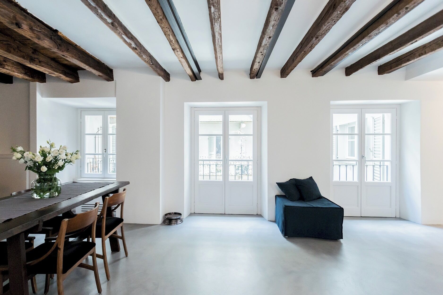 A spacious room with exposed wooden beams, a dining area with a dark table and chairs, and a dark blue sofa.