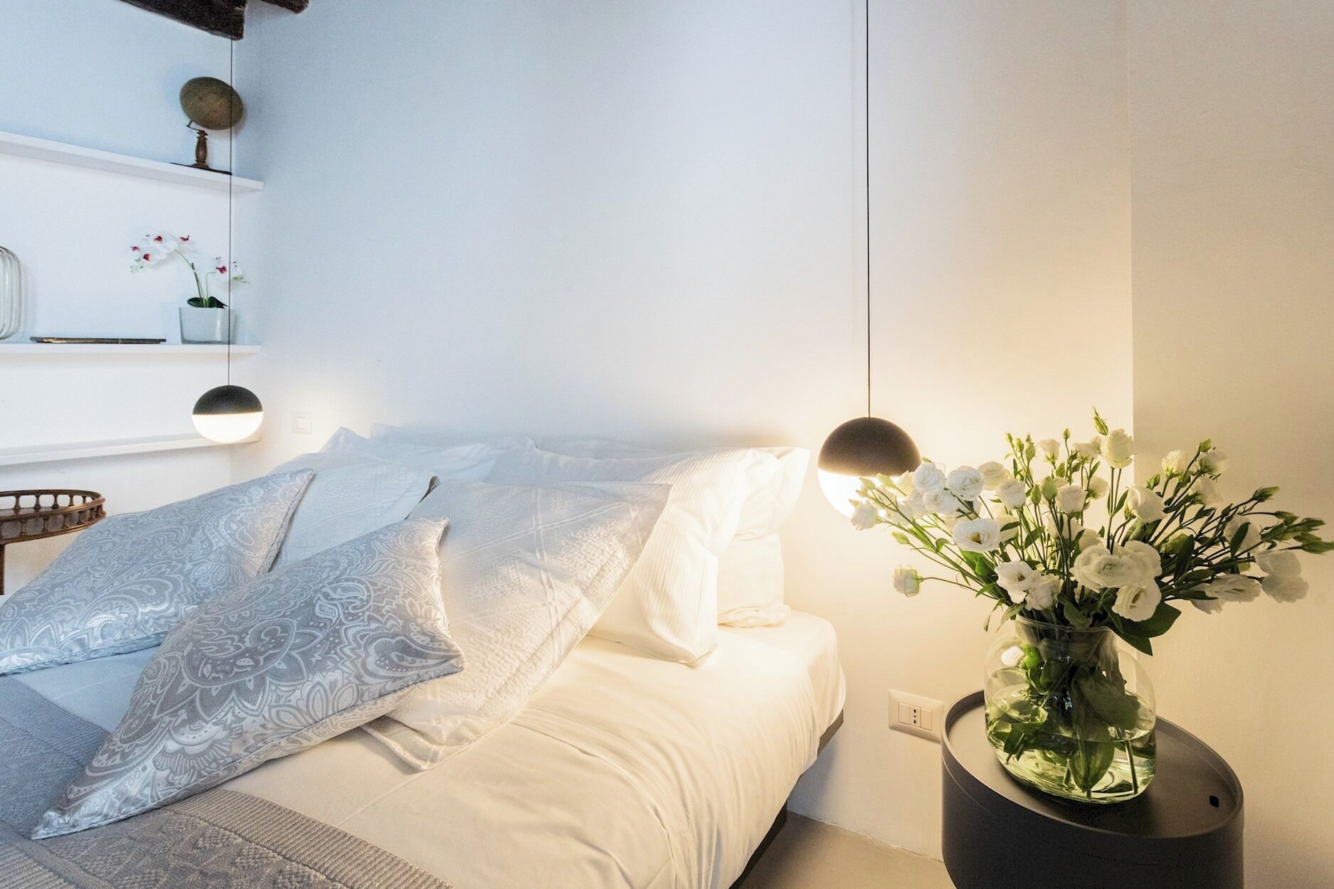 A neatly made bed with patterned pillows, a bedside table with a vase of flowers, and a wall-mounted shelf with decorative items.