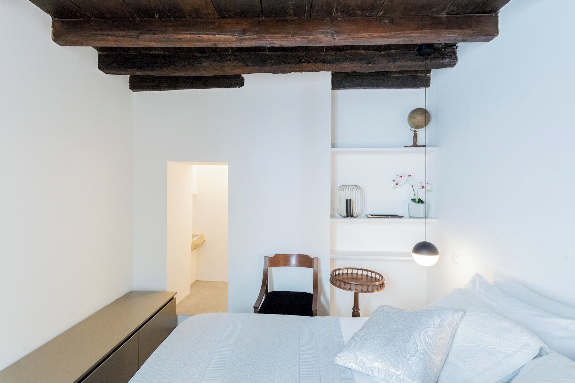 Ein Schlafzimmer mit einem Bett, einem Sessel, einem Regal mit Dekoration und einer Holzbalkendecke.