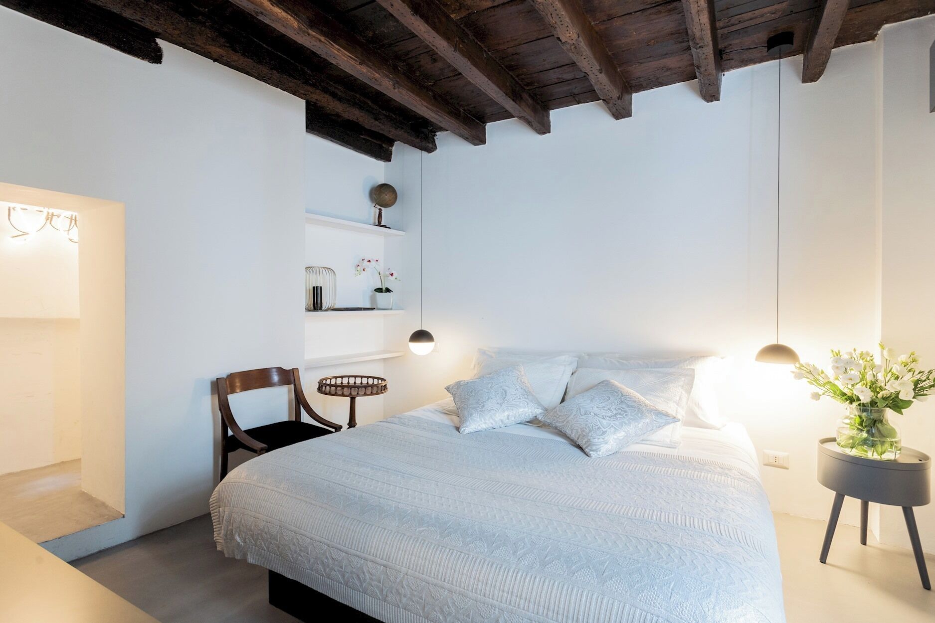 Ein Schlafzimmer mit einem großen Bett, Holzbalken an der Decke, einem Sessel, einem Nachttisch mit einer Pflanze und Regalen mit Dekoration.