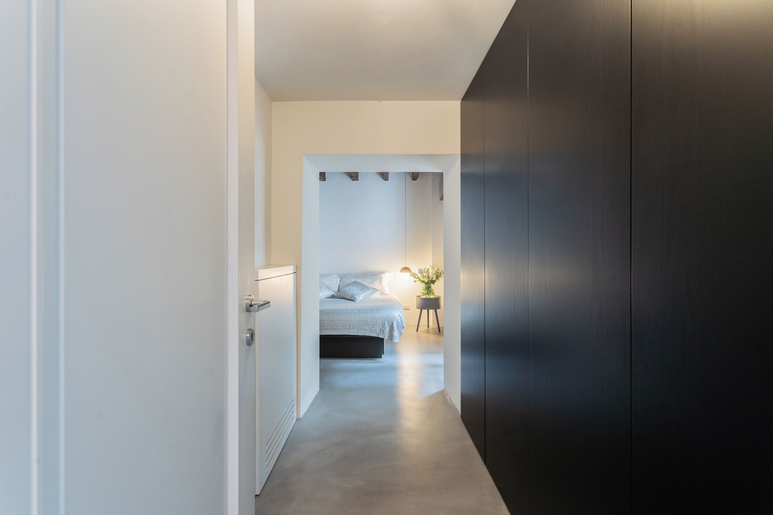 Een smalle gang die leidt naar een slaapkamer met een bed, nachtkastje en een lamp.