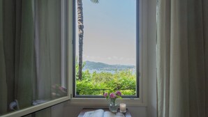 Une pièce avec une fenêtre donnant sur un paysage verdoyant, une petite table sur laquelle est posé un livre ouvert et un vase rempli de fleurs.