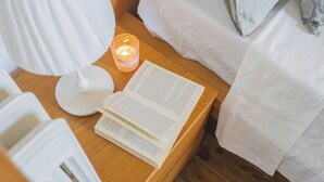 Un lit bien fait, avec du linge de lit blanc, une table de chevet sur laquelle repose un livre, une bougie allumée et une lampe blanche.