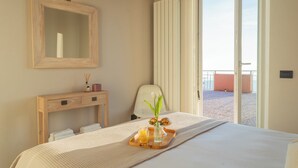 Ein Hotelzimmer mit einem Bett, einem Holztischchen mit Handtüchern, einem Spiegel, einer Vase mit Blumen und einem Tablett mit Getränken und Snacks.
