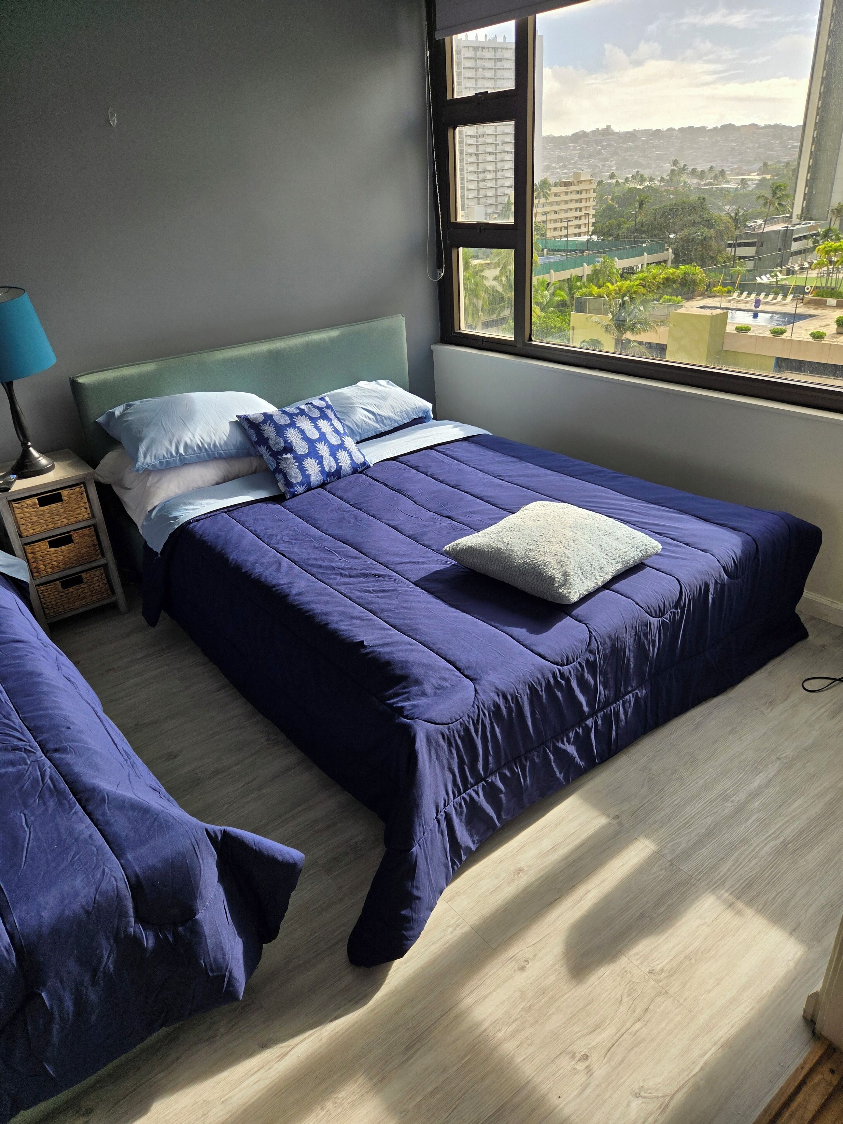Un dormitorio con cama, mesita de noche, lámpara y ventana con vista a edificios y árboles.