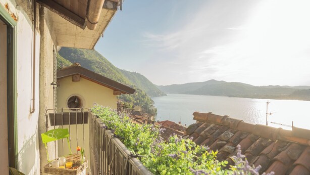 Un balcón con vistas a un lago y montañas, una pequeña mesa con una cesta y una flor, y un edificio con techo de tejas.