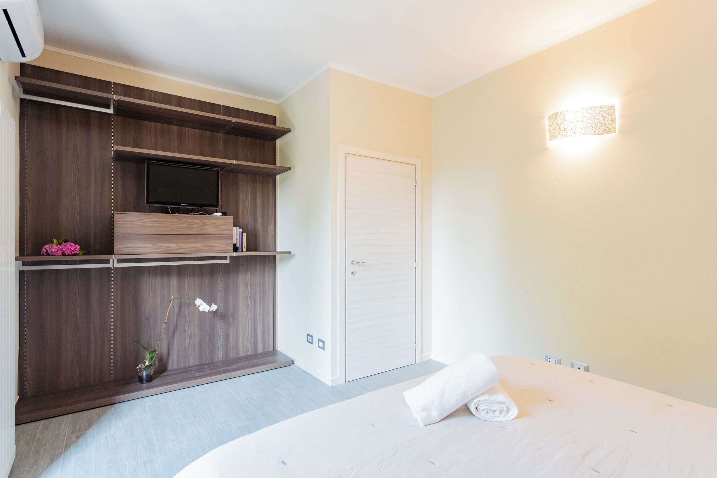 Habitación de hotel con cama, televisión en estante de madera, puerta blanca y lámpara de pared.