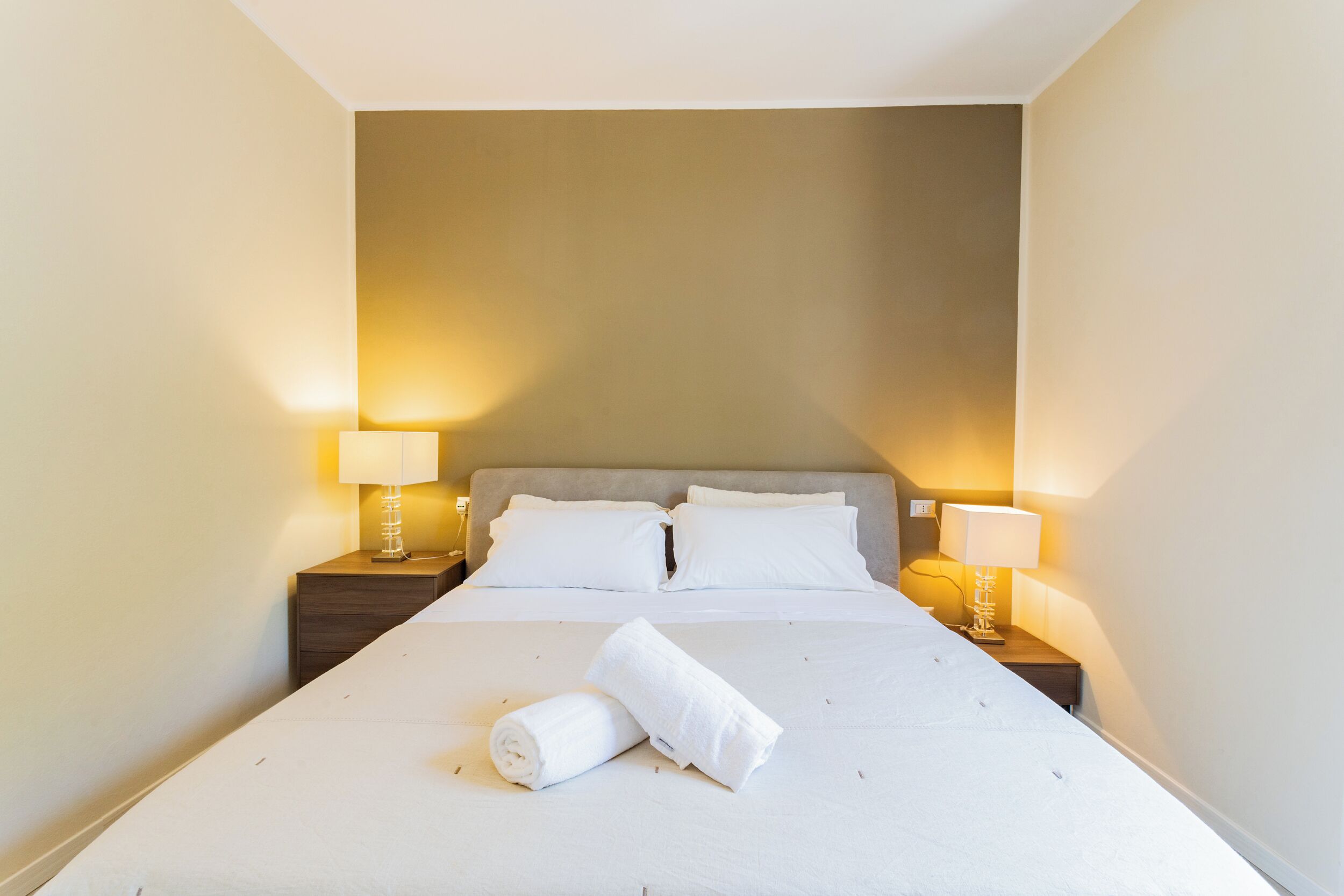 Een net opgemaakt bed met witte lakens en twee kussens, aan weerszijden met nachtkastjes en lampen, in een kamer met effen muren.