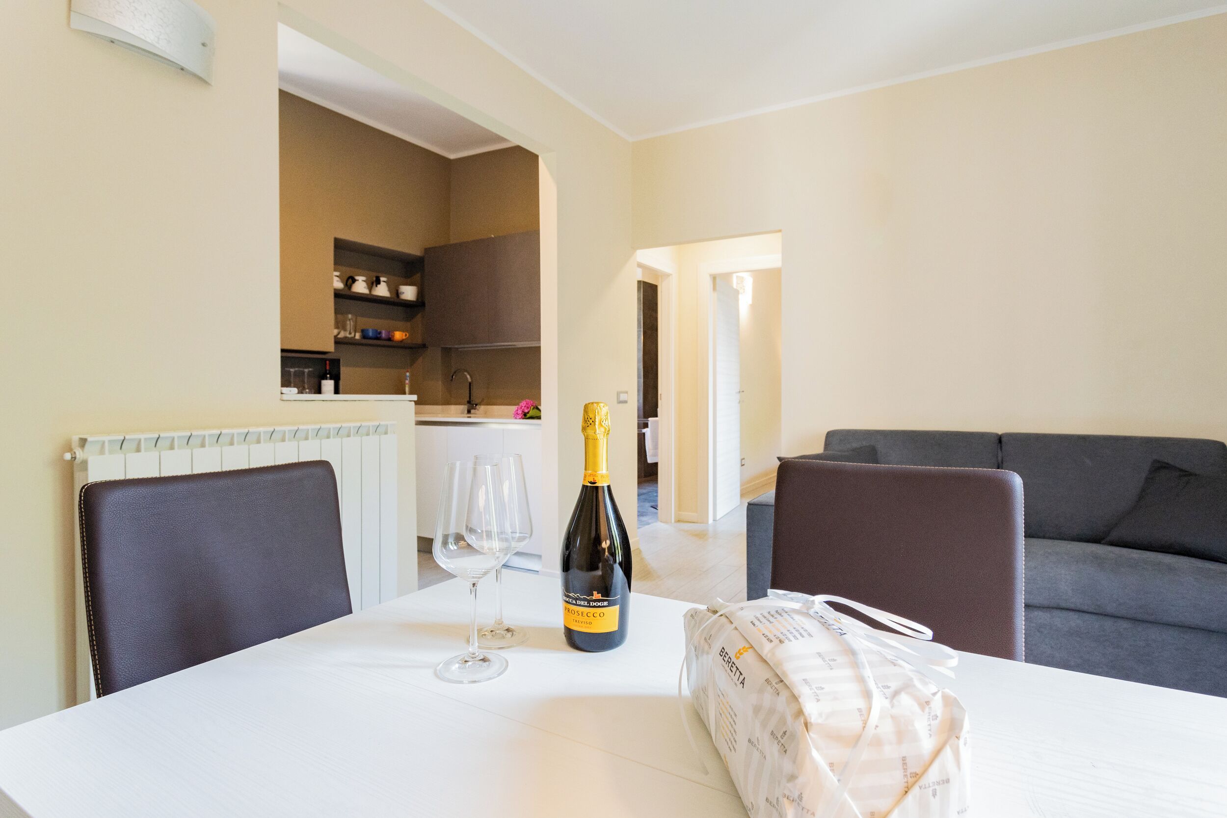 A dining table with a bottle of champagne, two wine glasses, and a bag of groceries in a modern living room with a grey sofa and a kitchen in the background.