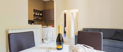 A dining table with a bottle of champagne, two wine glasses, and a bag of groceries in a modern living room with a grey sofa and a kitchen in the background.