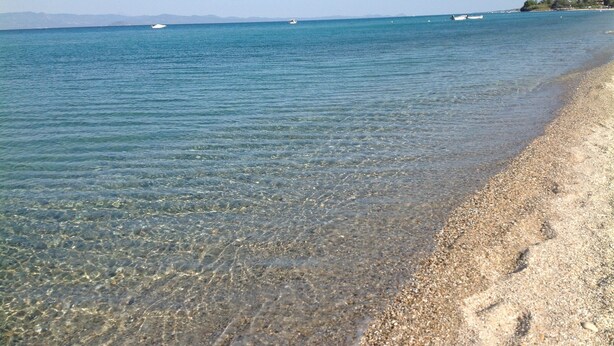 Una playa con aguas claras y poco profundas, una orilla arenosa y barcos a lo lejos.