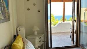 Un dormitorio con cama, almohadas, una mesita de noche, lámpara y vista al mar a través de una puerta abierta.