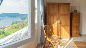 A room with a large window overlooking a lake and mountains, a wooden wardrobe, a wicker chair, and a small wooden table. - Top Lake View Villetta by Rent all Como (Cernobbio)
