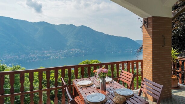 A wooden table set for two with plates, glasses, and a vase of flowers on a balcony overlooking a lake and mountains. - Top Lake View Villetta by Rent all Como (Cernobbio)