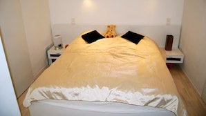En lille, pænt redt seng med et beige sengetæppe og to sorte puder, en bamse på sengen, og et natbord med en lampe og en lille højttaler.