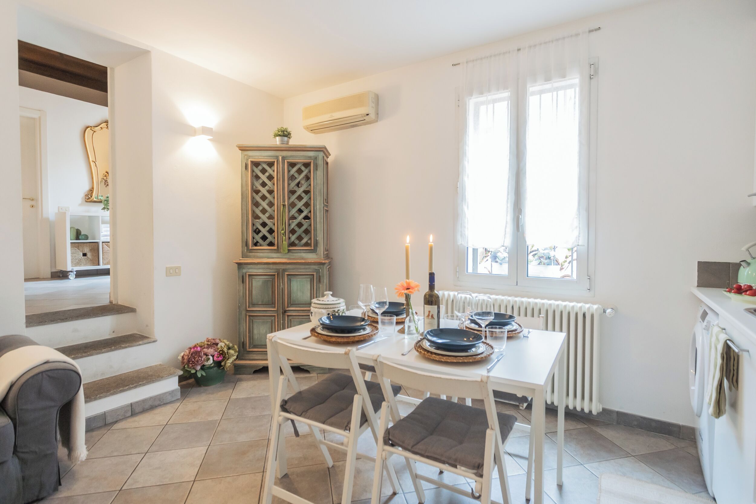 Un coin repas avec une table dressée pour deux personnes, un meuble avec un dessus décoratif et une fenêtre agrémentée de rideaux.