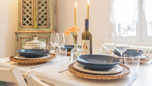 Une table à manger dressée avec des assiettes, des verres et des bougies, ainsi qu’une bouteille de vin et un vase avec une fleur.