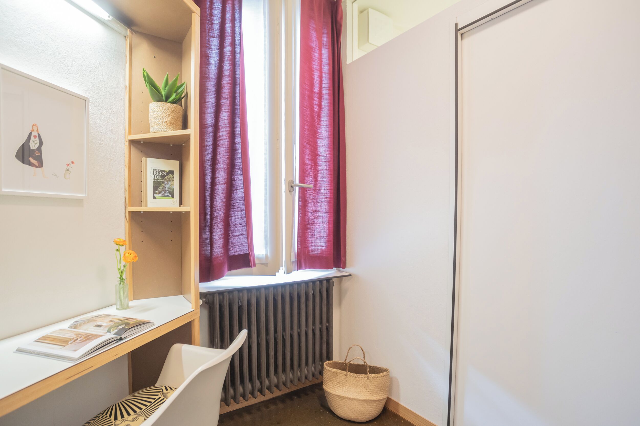 A small, neatly organized room with a desk, a chair, a radiator, a window with curtains, a shelf with a plant, and a basket.