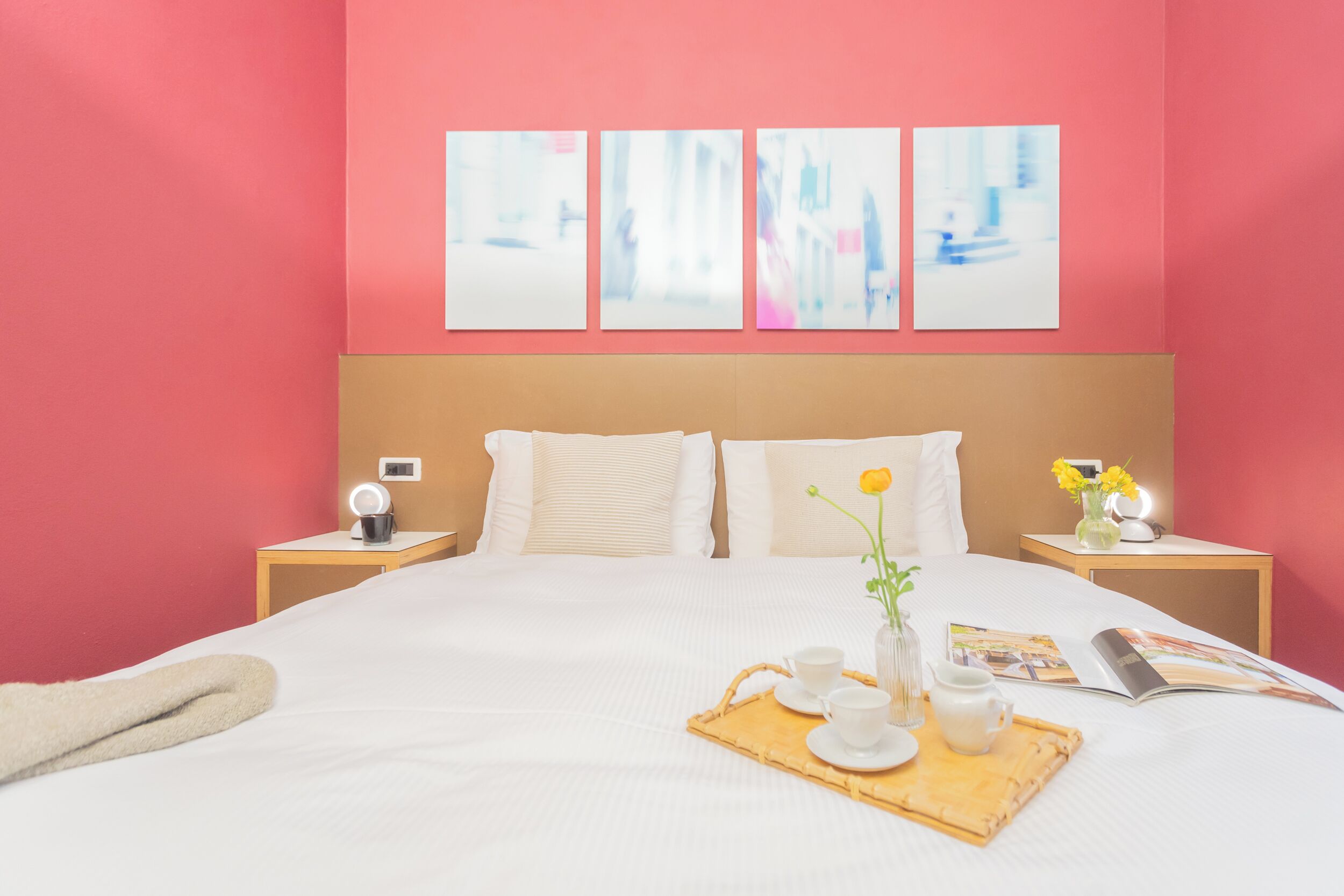 A hotel room with a large bed, white bedding, a bedside table with a lamp, a tray with cups and a vase of flowers, and a magazine on the bed.
