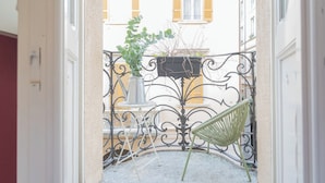A narrow outdoor corridor with a metal railing, a hanging planter with green foliage, and a green chair.