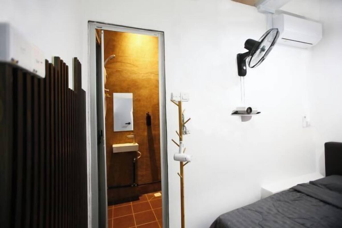 Quarto de hotel com cama, radiador, ventoinha na parede e porta para um banheiro.