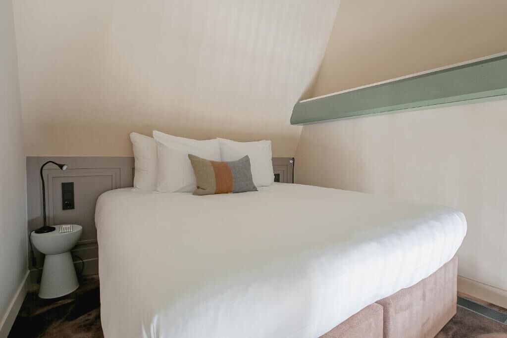 A neatly made bed with white bedding and a few pillows, a bedside table with a lamp, and a small white toilet.