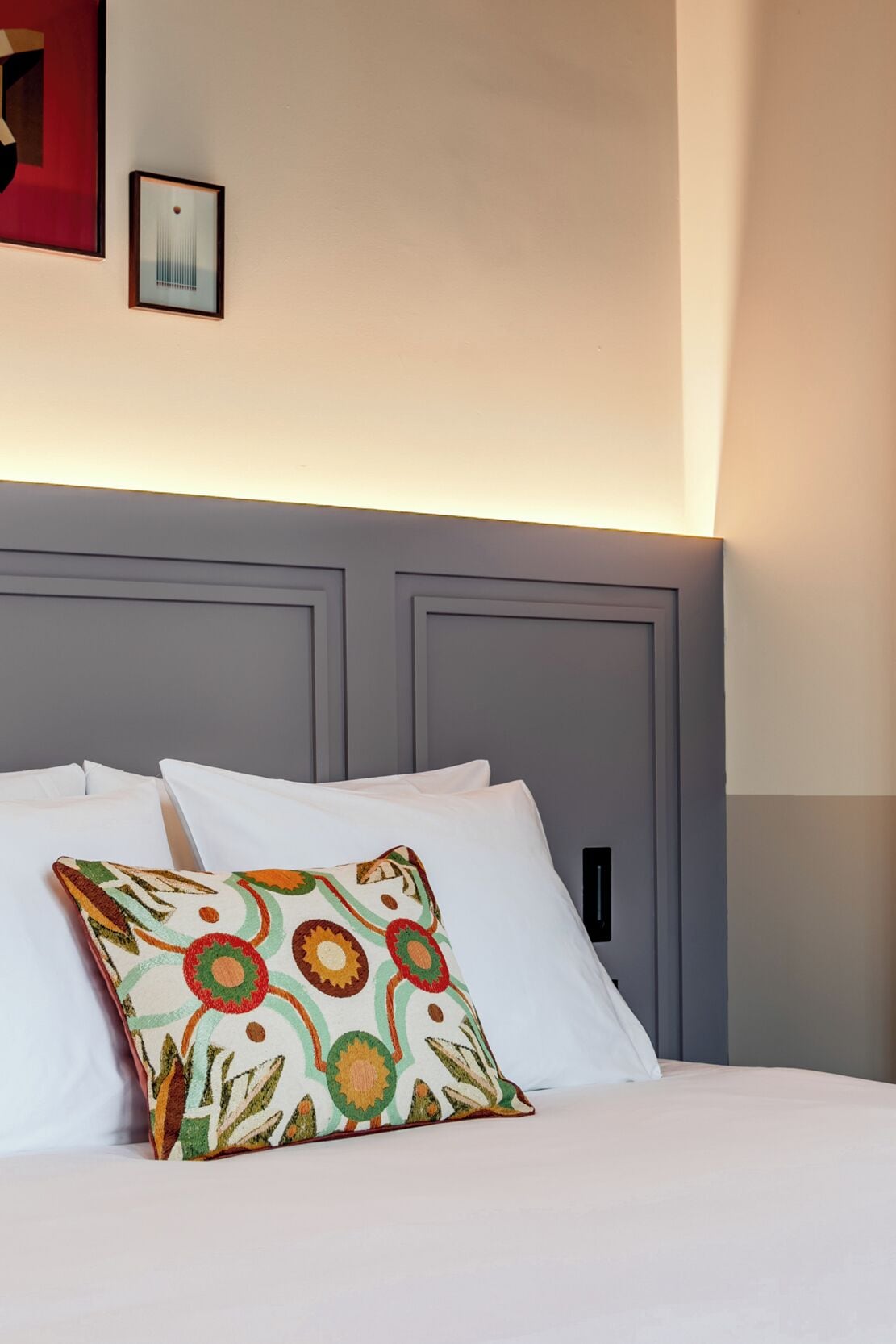 Una cama con sábanas blancas, un cojín de colores, un cabecero gris y un cuadro en la pared.