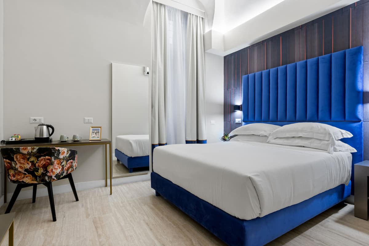 A modern hotel room with a large bed, a desk with a kettle, and a mirror.