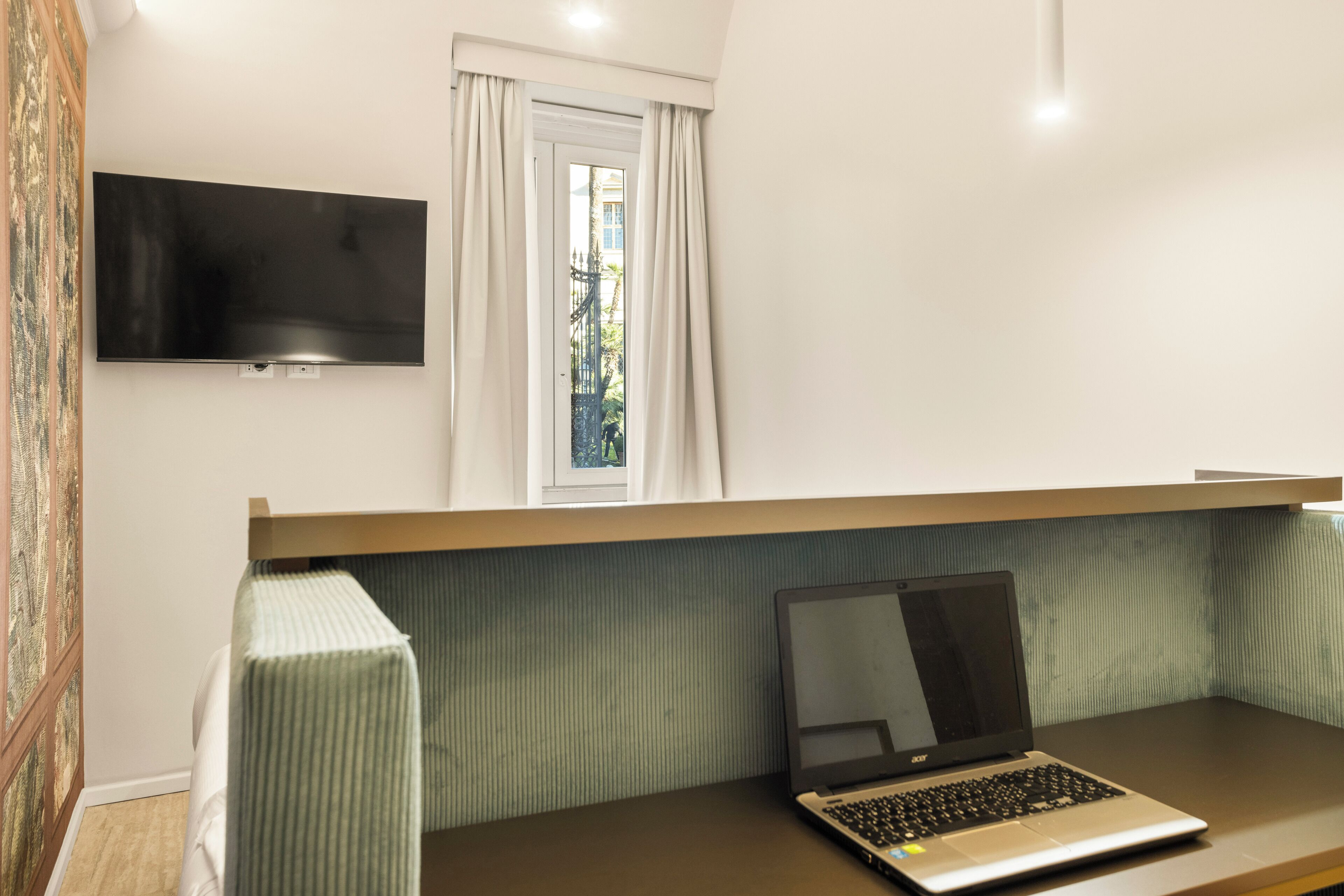 A bedroom with a bed, a laptop on a desk, a TV mounted on the wall, and a window with curtains.
