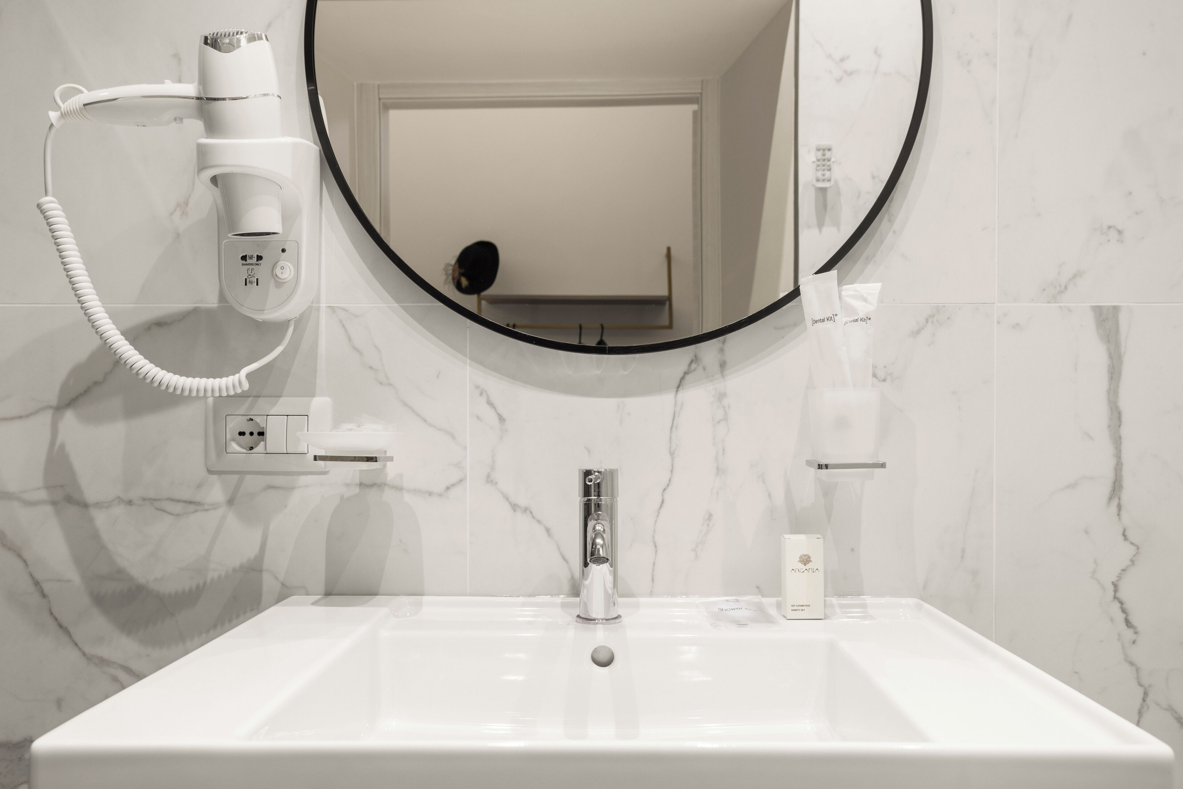 A bathroom with a white sink, a round mirror, a hairdryer, and a bottle of shampoo.