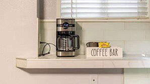 Una cocina con cafetera sobre una encimera blanca, un letrero de barra de café y una ventana con persianas.
