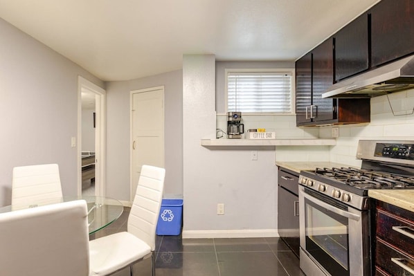A compact kitchen with a stove, oven, and coffee maker. There is a glass table with chairs and a recycling bin.
