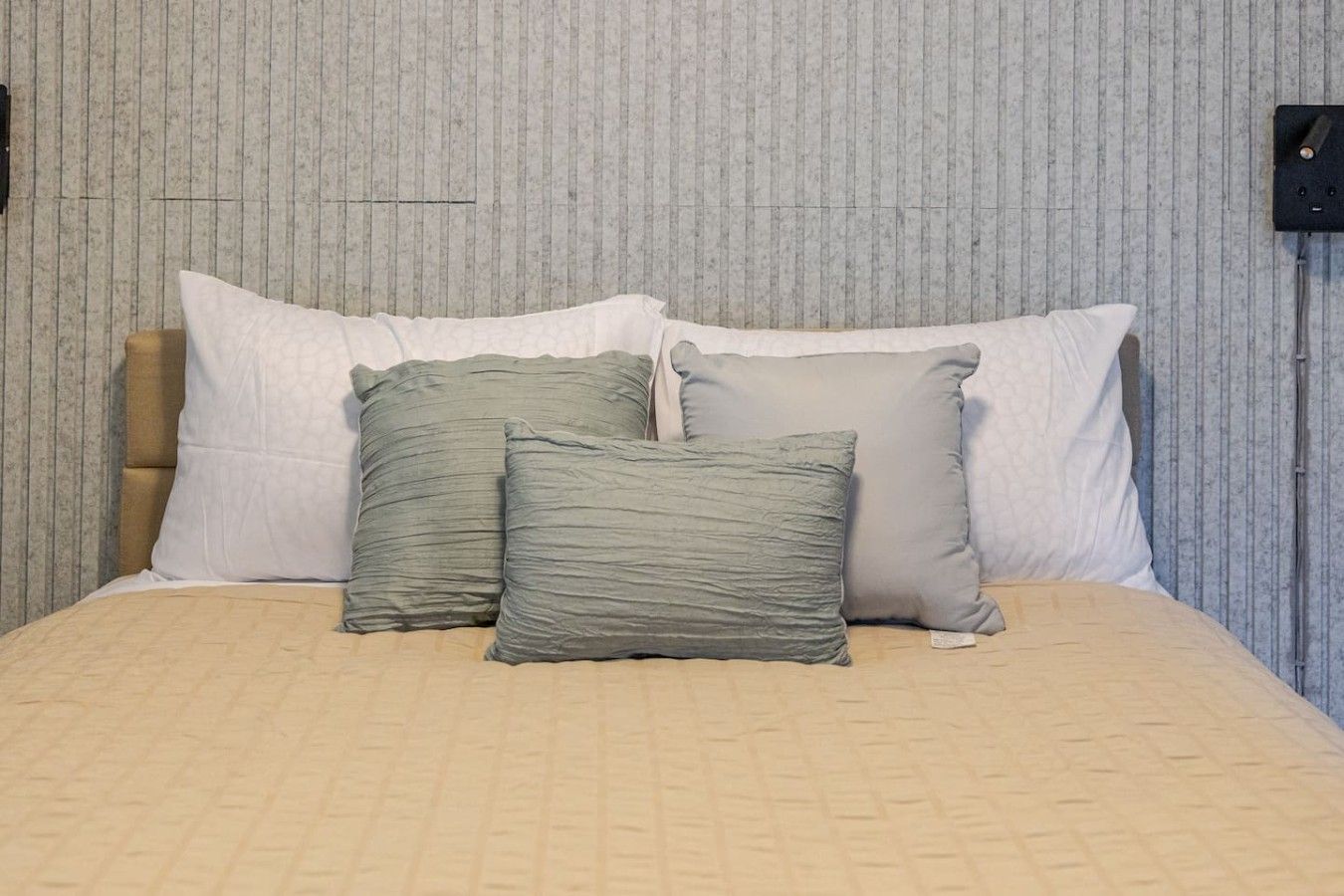 Ein Bett mit einer beigen Steudecke, zwei Kissen und einem Kopfteil vor einer strukturierten Wand.