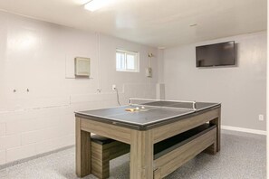 A room with a ping pong table, a television mounted on the wall, and a window providing natural light.